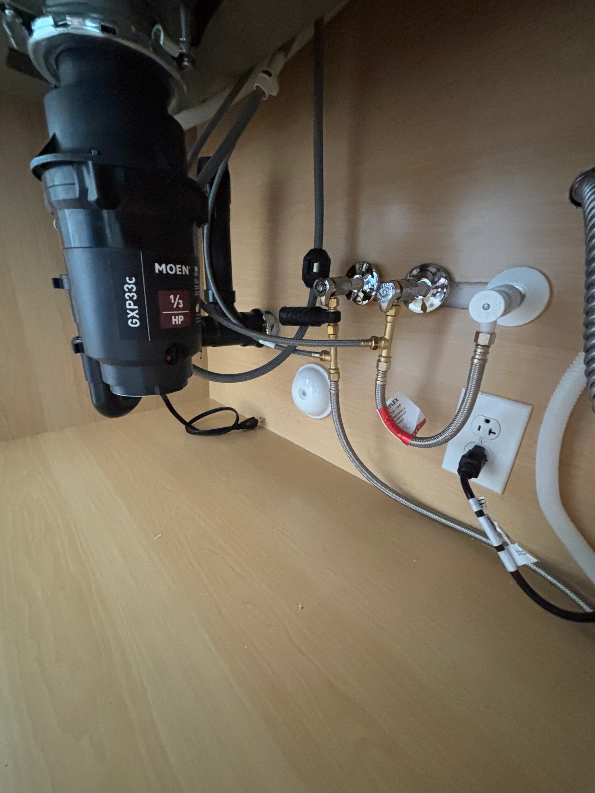 Moen 1/3 HP garbage disposal under a kitchen sink with chrome quarter-turn supply stops, braided faucet hoses, and the dishwasher outlet visible