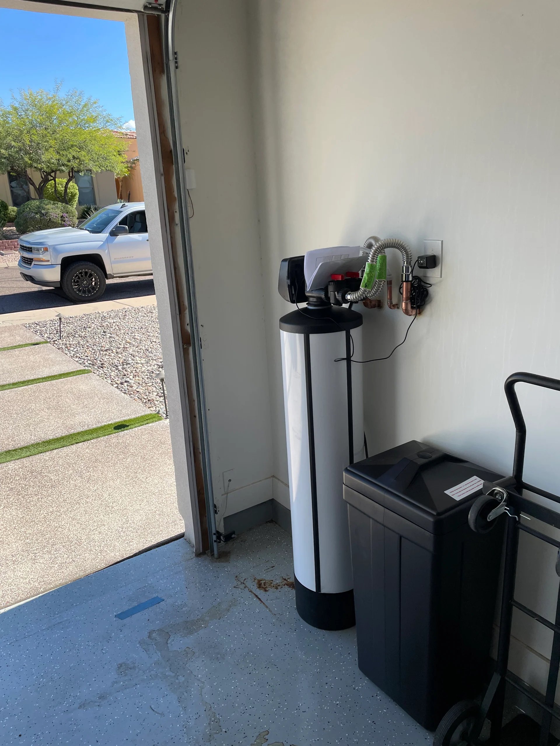 Whole-home water softener installed in a garage corner with copper supply lines and a service truck visible through the open door