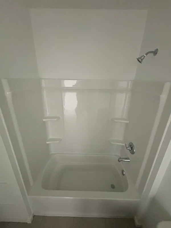 White acrylic alcove tub-and-shower combo with chrome showerhead, single-handle valve trim, and tub spout against gray tile