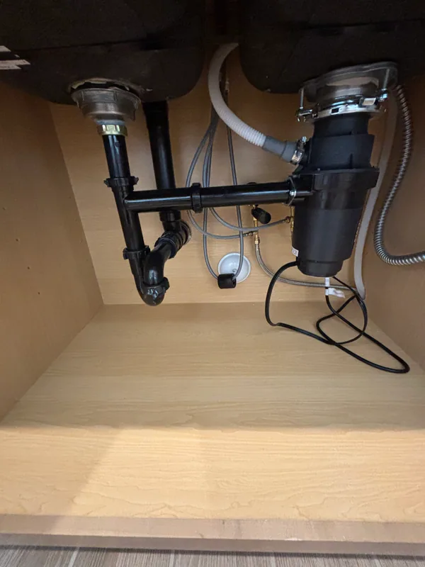 Garbage disposal under a kitchen sink with black ABS P-trap, dishwasher discharge loop, and braided supply lines inside a wood-veneer cabinet