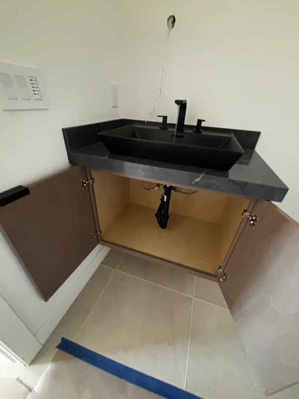 Charcoal-gray vanity with two matte-black wall-mount faucets above a deep dark basin, cabinet door open below to show the supply stops and braided lines inside