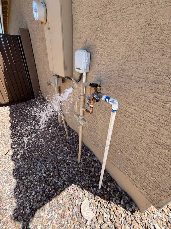 Water spraying from a PVC riser at an exterior stucco wall with a brass hose-bib and pressure gauge, soaking gravel in the side yard