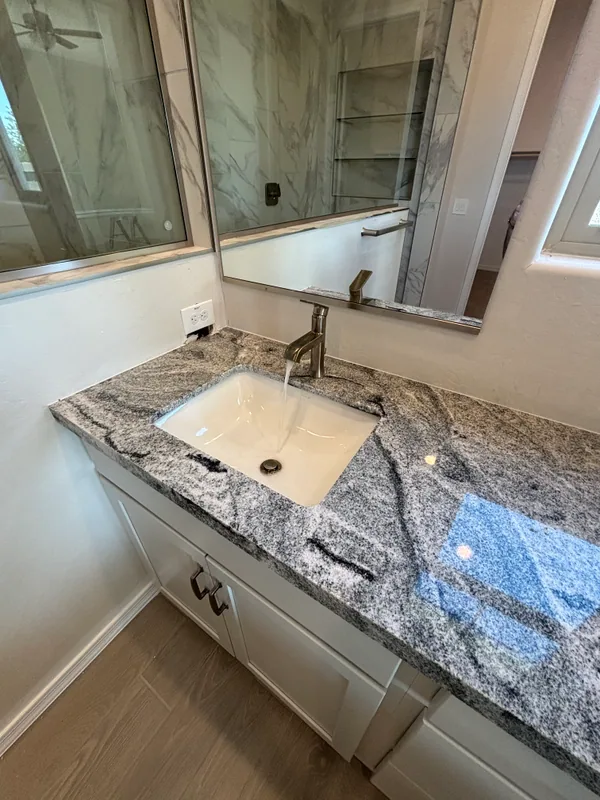 Undermount lavatory sink in a dark veined-granite counter with a brushed-bronze single-handle faucet, framed by a glass shower and tall window