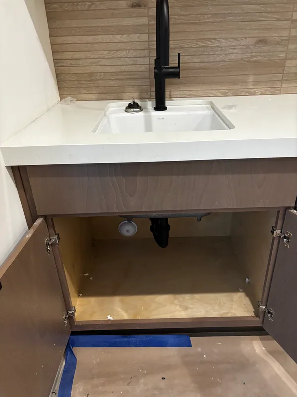 White undermount vanity sink with a matte-black single-handle wall-mount faucet on a wood-paneled wall, with the cabinet door open below to show the freshly run supply lines inside