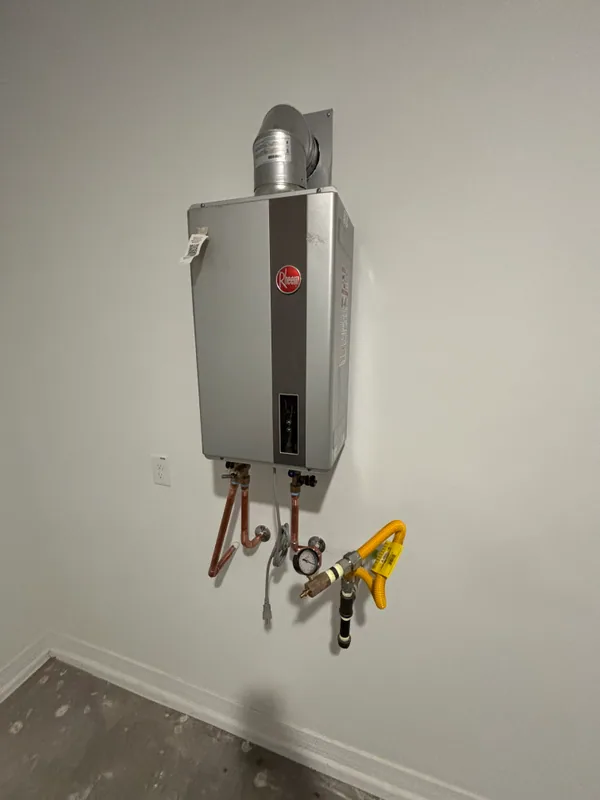 Rheem gas tank water heater mounted in a garage corner with copper supply lines, yellow flex gas connector, and PVC vent piping