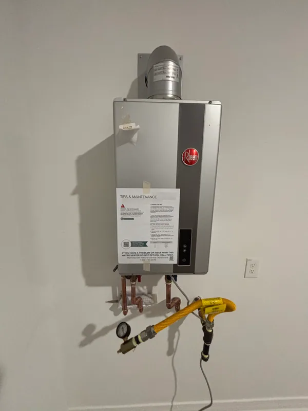 Rheem gas tank water heater on a utility wall with manufacturer tip-and-maintenance tag attached, copper supply lines, gauge, yellow flex gas connector, and PVC vent