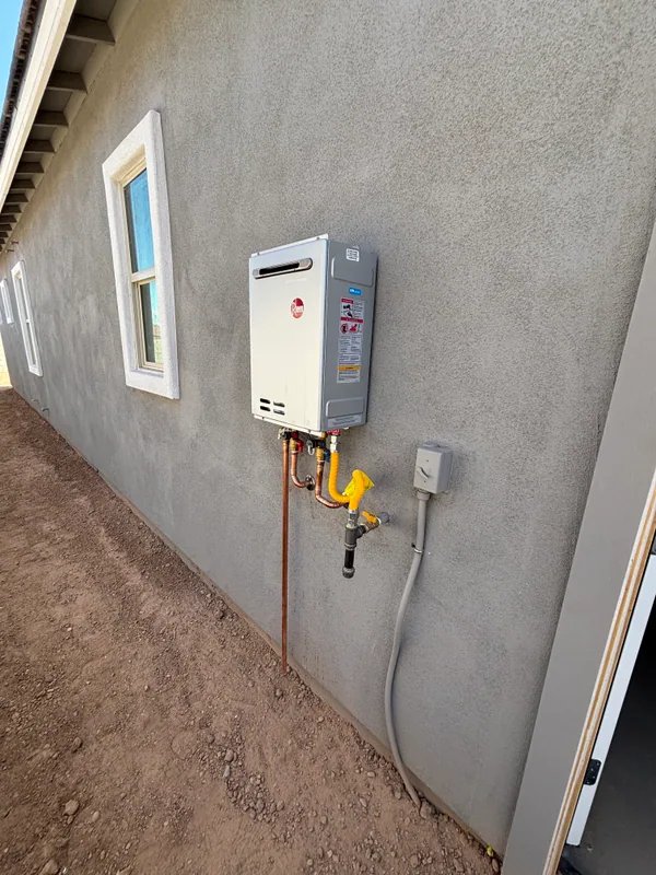 Rheem tankless water heater mounted on a tan stucco exterior wall with copper supply lines, gas shutoff, and yellow flex gas connector running to the meter