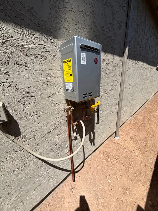 Rheem tankless water heater staged on a concrete driveway with copper supply lines, gas valve, and yellow flex connector ready for mounting