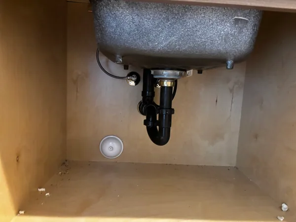 Under-sink view of a stainless basin with a freshly installed black P-trap, garbage disposal, and capped dishwasher outlet inside a plywood cabinet