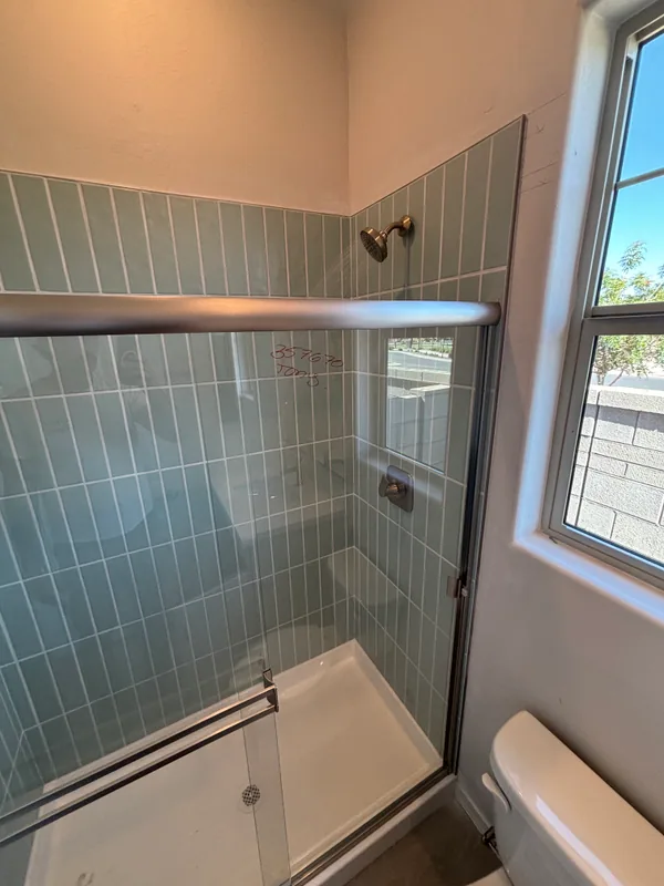 Tiled walk-in shower with light-blue subway tile, showerhead and valve trim on the back wall, and an aluminum sliding-door rail across the opening, with a small upper window above