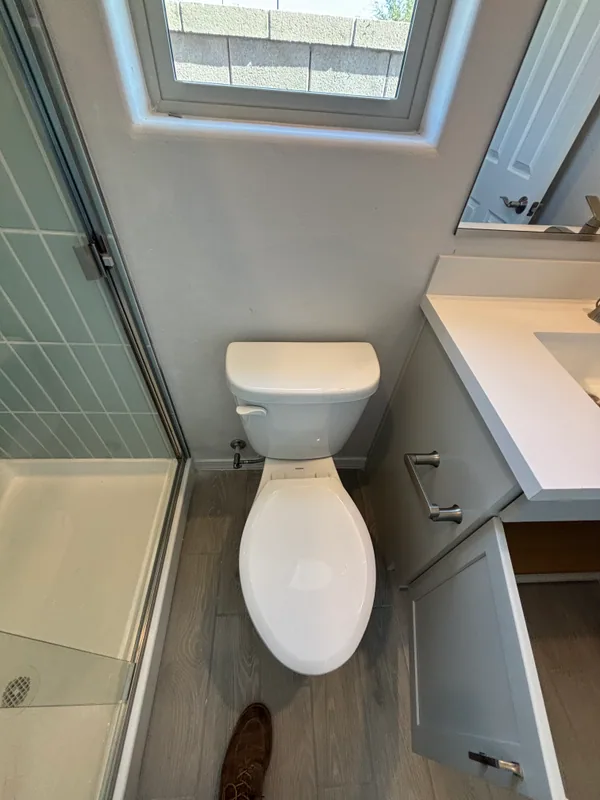 White elongated toilet installed on gray wood-look tile in a small bathroom with green subway-tile wainscot and a frosted window above