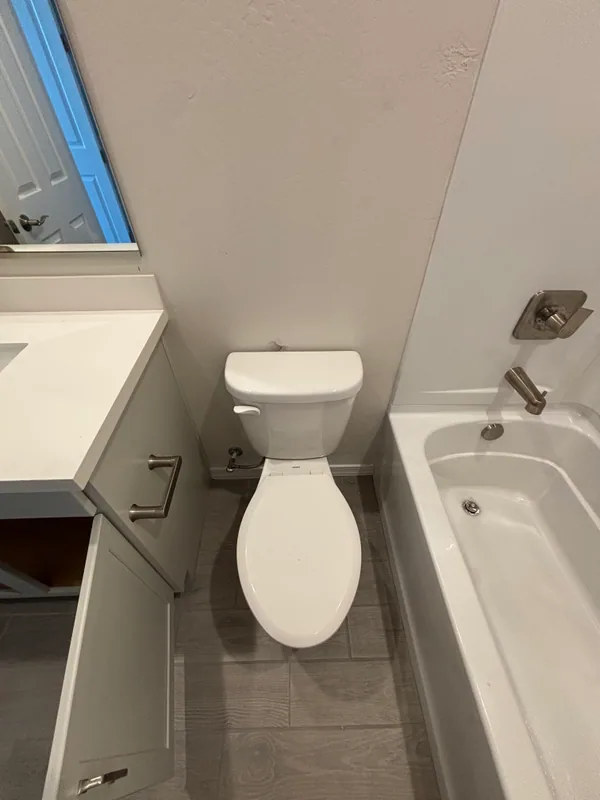 White elongated toilet set next to an alcove bathtub with brushed-nickel fixtures and tile surround, with a vanity visible behind