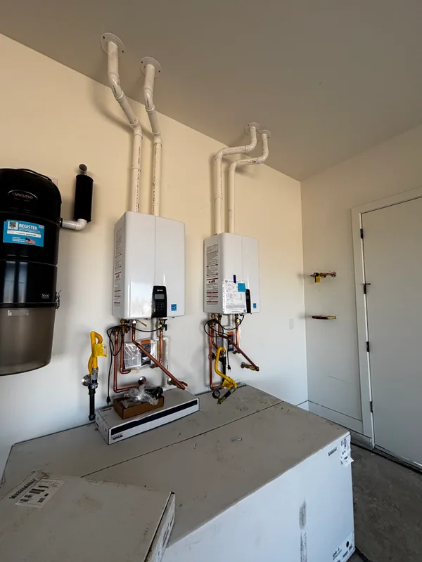 Two Rheem tankless water heaters mounted side-by-side on a garage wall with copper manifolds, yellow flex gas connectors, and a black water softener tank below