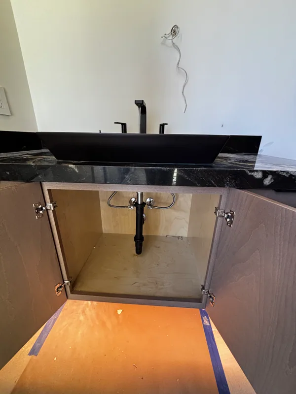 Vanity clad in dramatic veined black marble with two matte-black wall-mount faucets above, cabinet door open below to expose the matte-black valve body, copper supply, and braided supply hose inside