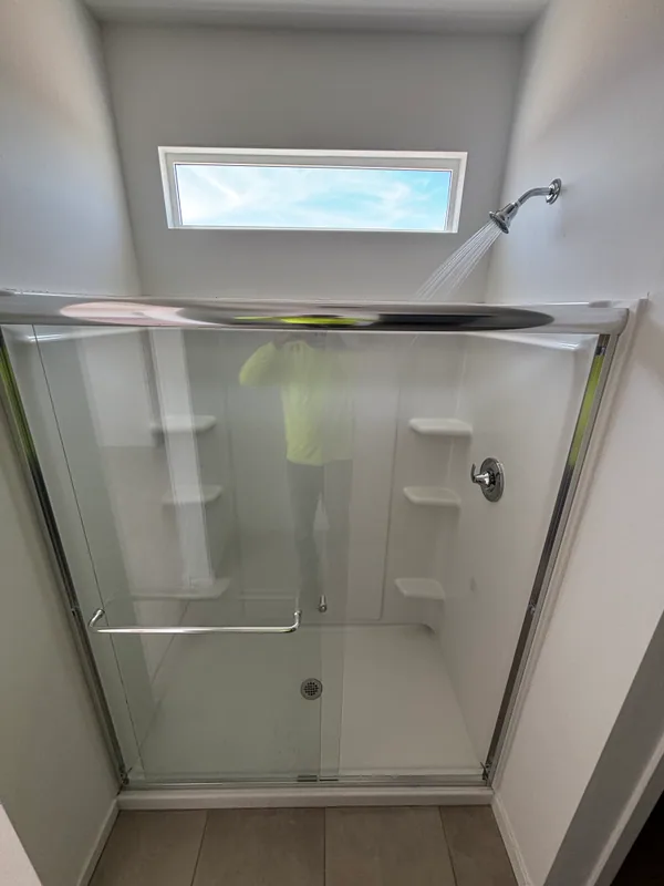 White acrylic walk-in shower with chrome sliding glass doors, fixed showerhead, and small window letting in daylight
