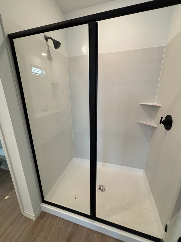White acrylic walk-in shower with matte-black framed glass enclosure, decorative vent panel, and built-in soap shelf