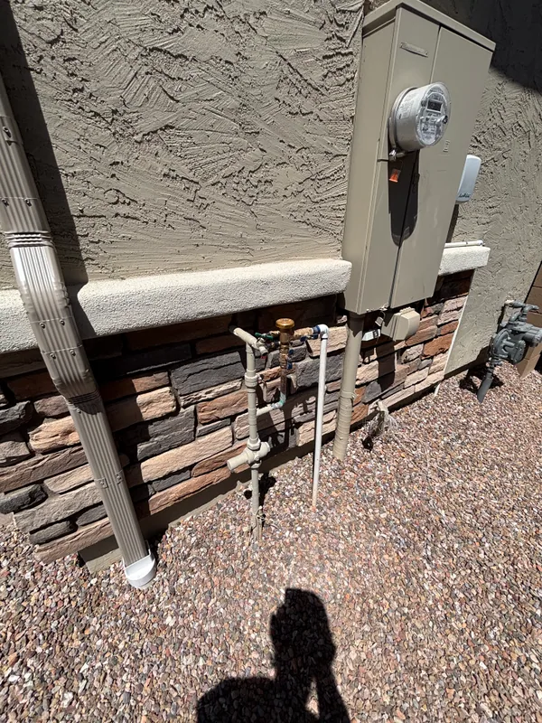 Water main stack — copper risers, brass shutoff, and backflow assembly — coming up out of fresh sand against a stacked-stone veneer column, with the home's electric meter pedestal on the left